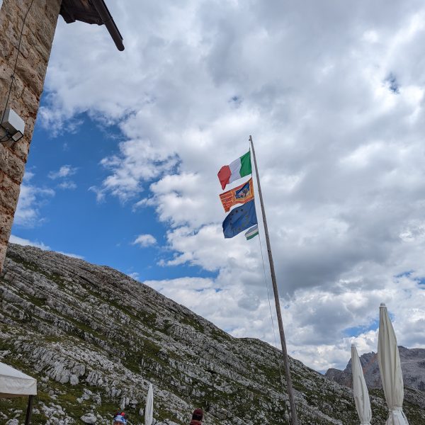 4 Flags flown on a pole next to an Italian Rifugio. The bottom most flag is smaller and it is the Ladin Flag