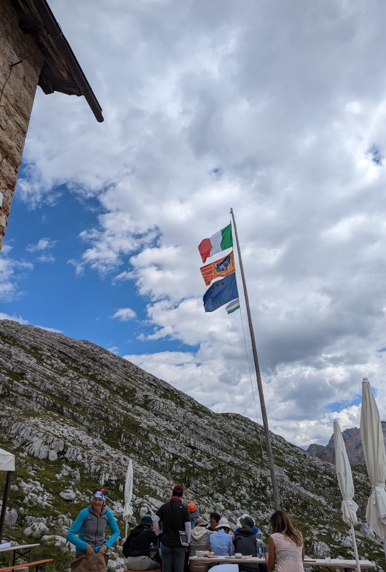 4 Flags flown on a pole next to an Italian Rifugio. The bottom most flag is smaller and it is the Ladin Flag